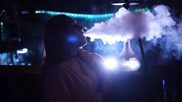 Close-up zijaanzicht van een meisje roken hookah op blauwe neon achtergrond. De media. Vrouw tijdens het roken van een hookah in een club, uitademen van een mist wolk en chillen. — Stockfoto