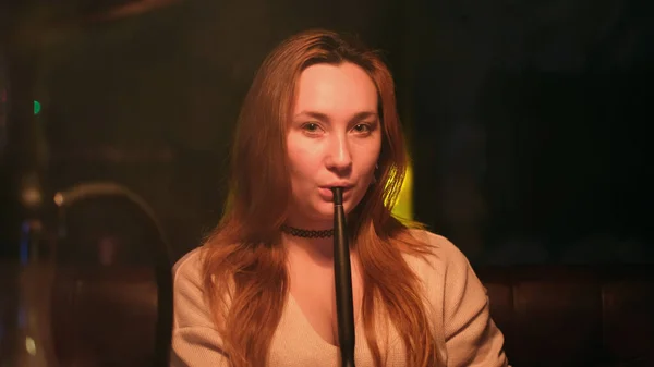 Menina de cabelos vermelhos sexy com decote profundo e exalando fumaça de narguilé direto para a câmera. Mídia. Mulher bonita passar o tempo enquanto fuma shisha. — Fotografia de Stock