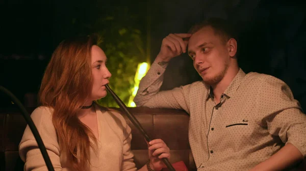Mladý milující pár kouří housku, zatímco sedí v kavárně u zelené neonové lampy. Média. Mladý manžel a manželka těší shisha a chatování. — Stock fotografie