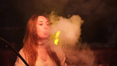 Kızıl saçlı seksi kız bir bar da açık kahverengi süveter giymiş koyu renk iç planda nargile içiyor. Medya. Güzel bayan neşeli görünüyor ve duman bulutları üflüyor.