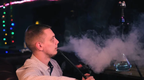 Jongeman is verslaafd aan een hookah. De media. Close-up zijaanzicht van een man in shirt uitademen dikke rook uit zijn mond om donkere achtergrond van cafe interieur. — Stockfoto