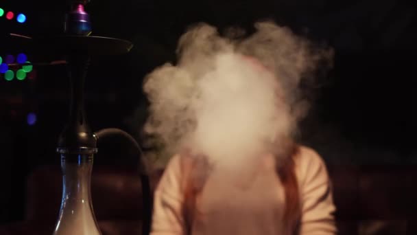 Νεαρή γυναίκα καπνίζει στο σκοτεινό μπαρ shisha lounge στο σκοτεινό φόντο του δωματίου. Μέσα ενημέρωσης. Γυναίκα με κοντά μαλλιά και μεγάλα μάτια καπνίζει ναργιλέ ενώ ξεκουράζεται σε εσωτερικούς χώρους, εκπνέοντας καπνό στην κάμερα. — Αρχείο Βίντεο