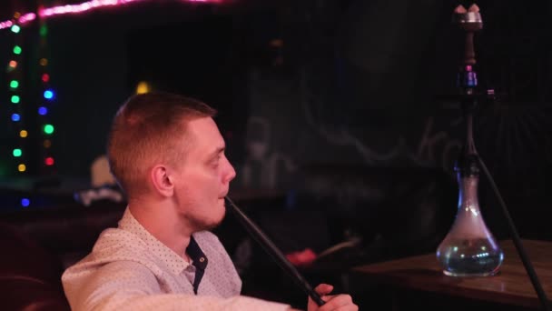 El joven es adicto a una cachimba. Medios. Vista lateral cercana de un hombre en camisa que exhala el humo espeso de su boca om fondo oscuro del interior de la cafetería. — Vídeos de Stock