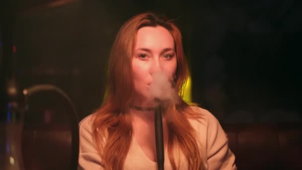 Рыжеволосая сексуальная девушка в лаундж-баре курит кальян, одетый в светло-коричневый свитер на темном внутреннем фоне. СМИ. Красивая леди игриво смотрит и дует облака дыма. — стоковое видео