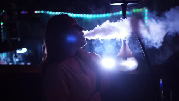 Gros plan vue de côté d'une fille fumant narguilé sur fond bleu néon. Les médias. Femme en fumant un narguilé dans un club, expirant un nuage de brouillard et se refroidissant. — Video