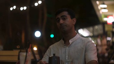 Genç adam sıcak çay ya da kahve içerken geceleri bir yazlık kafede yemek yiyor. Kavram. Kızgın siyah saçlı erkek, elinde kırmızı bardakta sıcak bir içecekle dışarıda birini bekliyor..