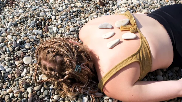 Μια νεαρή γυναίκα είναι ξαπλωμένη σε μια παραλία με βότσαλα. Έννοια. Άνδρας χέρι βάζοντας καυτές πέτρες στην πλάτη ενός νεαρού κοριτσιού με μαγιό με αφρόμπρους, το καλοκαίρι χαλαρώστε. — Φωτογραφία Αρχείου