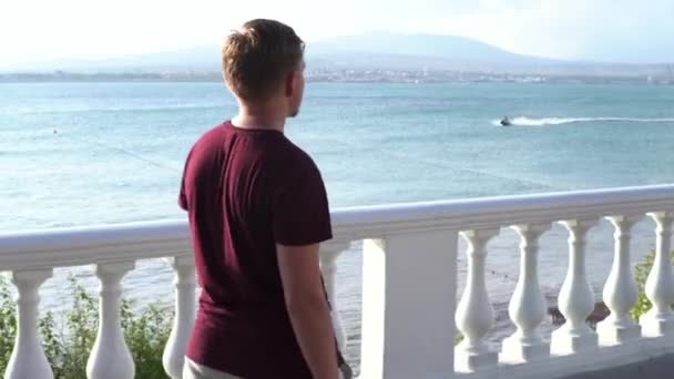 Blond man loopt langs de promenade in een zuidelijke stad aan zee. De media. Pensive jonge man wandelen langs de blauwe rimpel zee en bergen, concept van de zomer reis. — Stockvideo