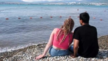 Deniz kenarında kırmızı şarap içen bir çift. Kavram. Saçları örülü romantik bir kadın ve sahilde otururken elinde kırmızı şarap bardakları olan bir adam..