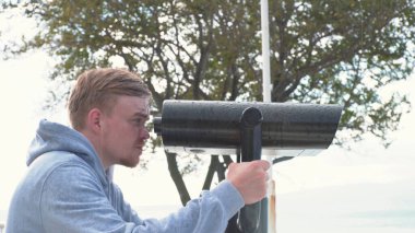 Genç adam gözlem güvertesindeki dürbünlerden bakıyor. Medya. İnsan yazın yağmurda dürbünle bakar. Adam, gözlem güvertesinde dürbünle bakan birini arıyor.