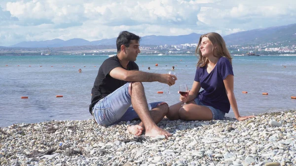 Ung man och kvinna slappna av sitter vid havet på solig dag. Media. Man och kvinna dricker vin ur glas som sitter på stranden. Semester med förälskade par till havs med glas vin — Stockfoto