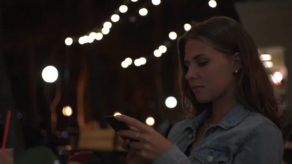 Beautiful woman reads message on background of lights of summer cafe. Media. Young woman looks at phone and smiles while sitting in street cafe in evening. Woman is waiting for date