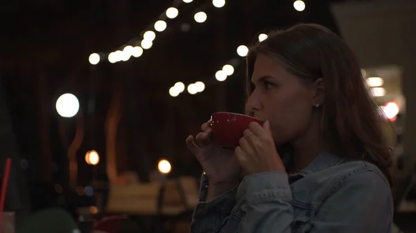 Beautiful woman reads message on background of lights of summer cafe. Media. Young woman looks at phone and smiles while sitting in street cafe in evening. Woman is waiting for date — Stock Photo, Image