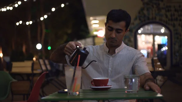 El hombre vierte agua hirviendo en la taza en el café. Medios. Atractivo hombre bebe café en la mesa cerca de la cafetería con luces de la noche. El hombre está esperando amada en la fecha en la cafetería al aire libre — Foto de Stock