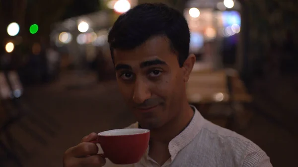 Člověk pije čaj a mrká. Média. Pohledný muž pije kávu a mrká na večerní světla. Muž flirtuje nad šálkem kávy v venkovní kavárně ve večerních hodinách — Stock fotografie