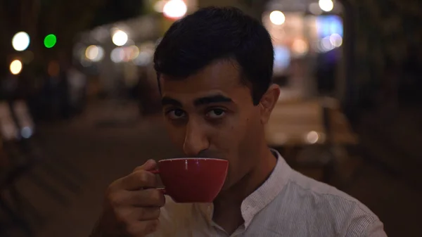 Hij drinkt thee en knipootjes. De media. Knappe man drinkt koffie en knipoogt op achtergrondverlichting. Man flirt over kopje koffie in de openlucht in cafe in de avond — Stockfoto