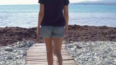 Deniz ufkunun arka planında sahilde yürüyen bir kadın. Medya. Güneşli bir günde tahta patika boyunca yürüyen şortlu genç bir kadının arka görüntüsü. Denizde harika bir tatil