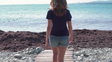 Deniz ufkunun arka planında sahilde yürüyen bir kadın. Medya. Güneşli bir günde tahta patika boyunca yürüyen şortlu genç bir kadının arka görüntüsü. Denizde harika bir tatil