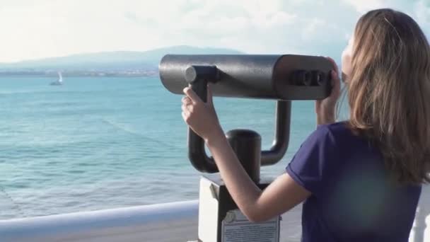 Piękna kobieta patrząca przez lornetkę na tle morza i gór. Media. Młoda kobieta ogląda krajobraz morski z górami w parę lornetek w słoneczny dzień — Wideo stockowe