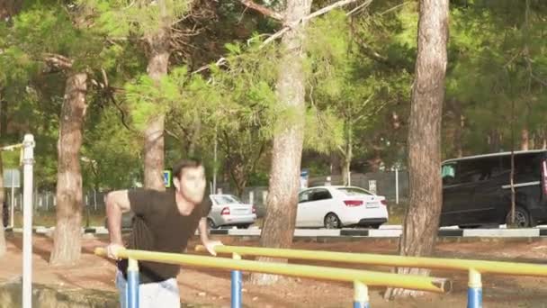 Joven haciendo flexiones en barras irregulares. Medios. Hombre joven entrena en el terreno de juego con push-UPS en barras irregulares. Entrenamiento con press de banca al aire libre en un día soleado — Vídeos de Stock