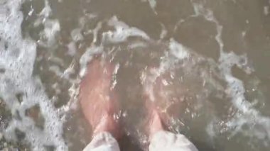 Sahilde dalgalarla ayaklar. Medya. Kayalık sahilde dalgalı deniz dalgalarıyla serinleyen erkek ayaklarının yakın çekimi. Deniz dalgalarıyla sahilde ayaklar