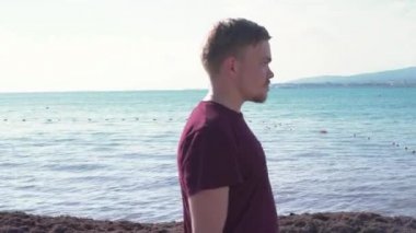 Genç adam güneşli bir günde denizin arka planında yürür. Medya. Genç adam güneşli bir günde güzel deniz kıyısında yürür. Adam deniz manzarasının arka planında yürüyor dağlarla ve kasabayla