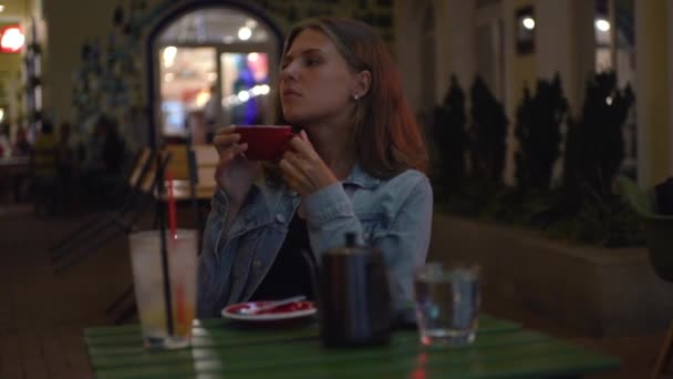 Красива жінка п'є каву за вуличним столиком ввечері. Медіа. Жінка п'є каву на відкритому столі ввечері. Затишний літній вечір з чашкою кави в кафе з вогнями — стокове відео