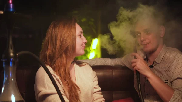 Cute couple in love relaxing on a date. Media. Caucasian young man and woman in black smoking a hookah while sitting at the table in cafe indoors together. — Stock Photo, Image