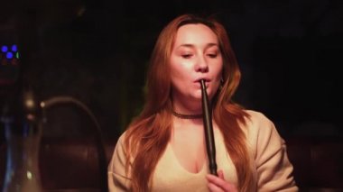Koyu yakalı ve nargile dumanı doğrudan kameraya üfleyen seksi kızıl saçlı kız. Medya. Güzel bir kadın sigara içerken vakit geçiriyor..