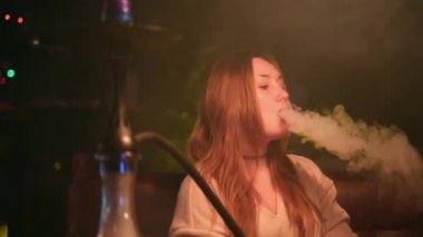 Çekici genç bir kadın restoranda nargile içiyor. Medya. Zarif bir kadın ve boynu bükük bir model. Kafede nargileyle oturuyor ve duman bulutu yaratıyor..