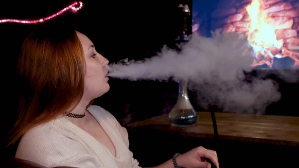 Sidovy över en rödhårig flicka som röker shisha framför TV-skärmen med brinnande eld. Media. Ung vacker kvinna med en svart choker runt halsen rökning hora på bakgrunden av falska — Stockfoto