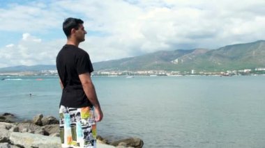 Erkek turist kayanın üzerinde duruyor ve okyanusun tadını çıkarıyor. Kavram. Kayalıklarda duran bir adamın arka görüntüsü ve masmavi denize bakan bir adam, dağlar, bulutlu gökyüzü ve kıyı kasabası..