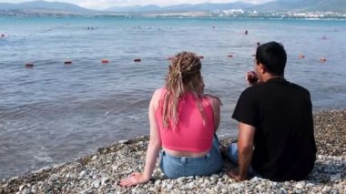 Deniz kenarında kırmızı şarap içen bir çift. Kavram. Saçları örülü romantik bir kadın ve sahilde otururken elinde kırmızı şarap bardakları olan bir adam..