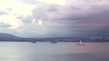 Yelkenlilerle göz kamaştırıcı mor gün batımı manzarası. Medya. Yüzeyinde yelkenli yatları, dağları, bulutlu leylakları ve pembe bulutları olan doğal deniz manzarası.