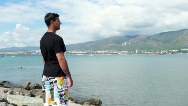 Muž turista stojící na skále a užívající si oceán. Koncept. Pohled zezadu na muže stojícího na skalním útesu a obráceného k azurovému moři, horám, zatažené obloze a pobřežnímu městu. — Stock video