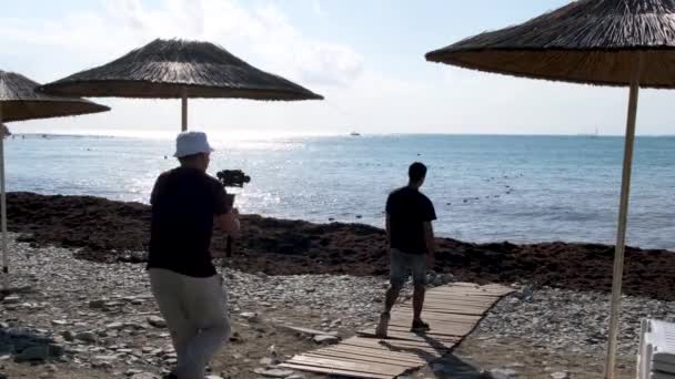 Videógrafo tirando fotos e vídeos das vistas do mar e um homem vindo para a água. Conceito. Visão traseira de um homem com uma câmera filmando outro cara na praia. — Vídeo de Stock