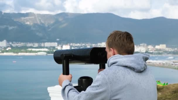 Jongeman kijkt door een verrekijker in de regen. Concept. De mens kijkt door verrekijker op observatiedek op de achtergrond van de zeekust bij regenachtig weer — Stockvideo