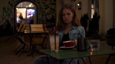 Kafede bir fincan kahve ve masada bir kokteyl ile yalnız başına oturan ve telefonu kullanan güzel, yalnız bir kızın portresi. Medya. Genç kız akşam restoranda sıkılmış görünüyor.