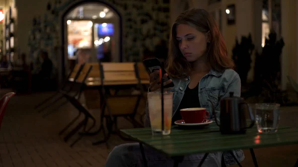 Kafede bir fincan kahve ve masada bir kokteyl ile yalnız başına oturan ve telefonu kullanan güzel, yalnız bir kızın portresi. Medya. Genç kız akşam restoranda sıkılmış görünüyor. — Stok fotoğraf