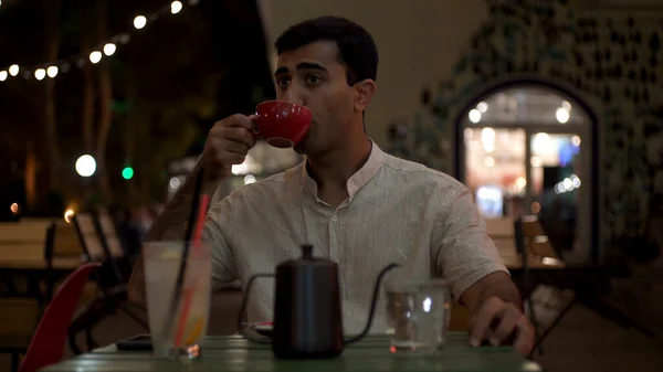 Egy fiatalember forró teát vagy kávét iszik, miközben egy nyári kávézó asztalánál ül esténként. A média. Ideges fekete hajú férfi vár valakire a szabadtéri teraszon egy forró itallal egy piros csészében.. — Stock Fotó
