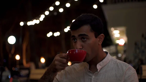 슬프게도 로맨틱 한 한 남자가 야외 식당에서 여자 친구를 기다리고 있습니다. 미디어. 흰색 셔츠를 입고 저녁늦게 혼자 앉아서 빨간 컵에서 뜨거운 음료를 마시는 지루 한 사람의 모습. — 스톡 사진
