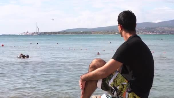 Muž cestující sedí na útesu nad mořem a dívá se daleko v dálce. Média. Boční pohled na mladého muže v jasných kraťasech a černé tričko těší letní dovolenou. — Stock video