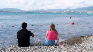 Güzel bir denizin, dağların ve arka planda bulutlu gökyüzünün keyfini çıkaran sevgi dolu bir çiftin arka görüntüsü. Kavram. Sahilde romantik bir buluşma, deniz kıyısında birlikte oturan kadın ve erkek..