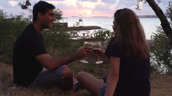 Älskade glada par dricker rött vin nära havet stranden under solnedgången tid. Media. Man och kvinna chattar medan du sitter på marken nära grönt gräs och buskar. — Stockfoto