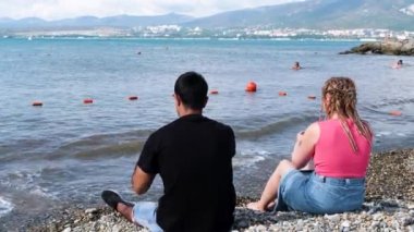 Güzel bir denizin, dağların ve arka planda bulutlu gökyüzünün keyfini çıkaran sevgi dolu bir çiftin arka görüntüsü. Kavram. Sahilde romantik bir buluşma, deniz kıyısında birlikte oturan kadın ve erkek..