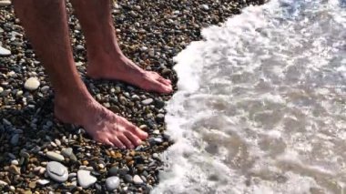 Çıplak ayaklı bir erkek, deniz tarafından yıkanan çakıl taşı plajında duruyor. Kavram. İnsan bacakları ve ayakları suyun altında, çakıl taşları şeffaf dalgalar aracılığıyla görülebilir..