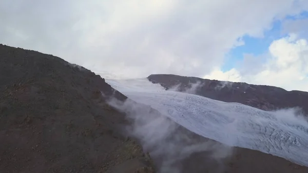 Amazing landscape with a mountain slope covered by cold ground and white snow. Clip. Aerial view of clouds flowing slowly, sliding up the hill on blue cloudy sky background.