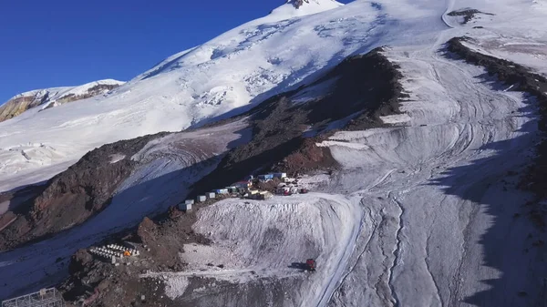A row of houses built on a snowy mountain slope on blue sky background. Clip. Aerial view of garage buildings and machines located high in the mountains.