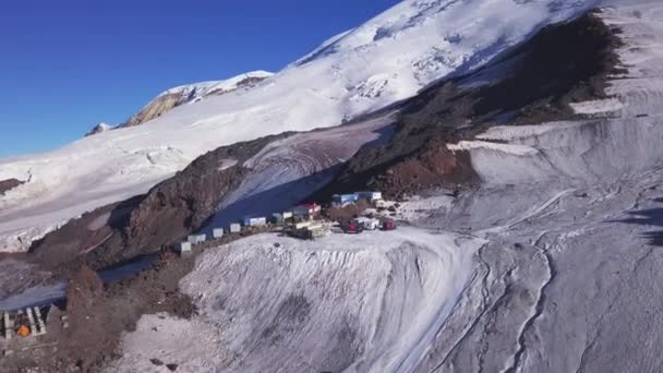 Uma fileira de casas construídas em uma encosta de montanha nevada no fundo do céu azul. Clipe. Vista aérea de edifícios de garagem e máquinas localizadas no alto das montanhas. — Vídeo de Stock