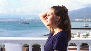 Kız deniz kıyısındaki yaz tatilinin tadını çıkarıyor. Medya. Mavi deniz arka planında mavi bir tişörtle şehrin seti boyunca yürüyen güzel bir kadının yan görüntüsü..